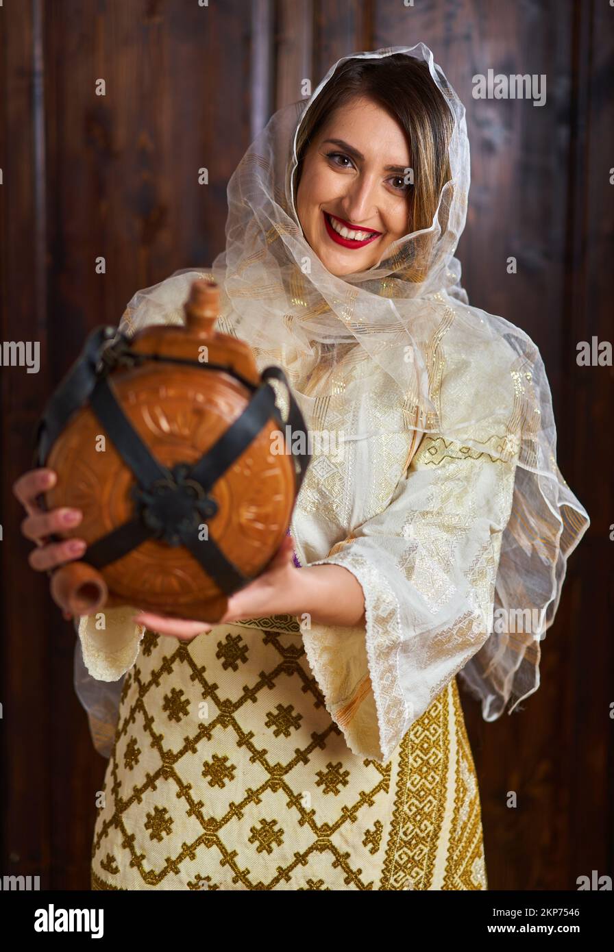 Giovane donna rumena in costume tradizionale sposa popolare in una casa d'epoca con una bottiglia di vino in legno Foto Stock