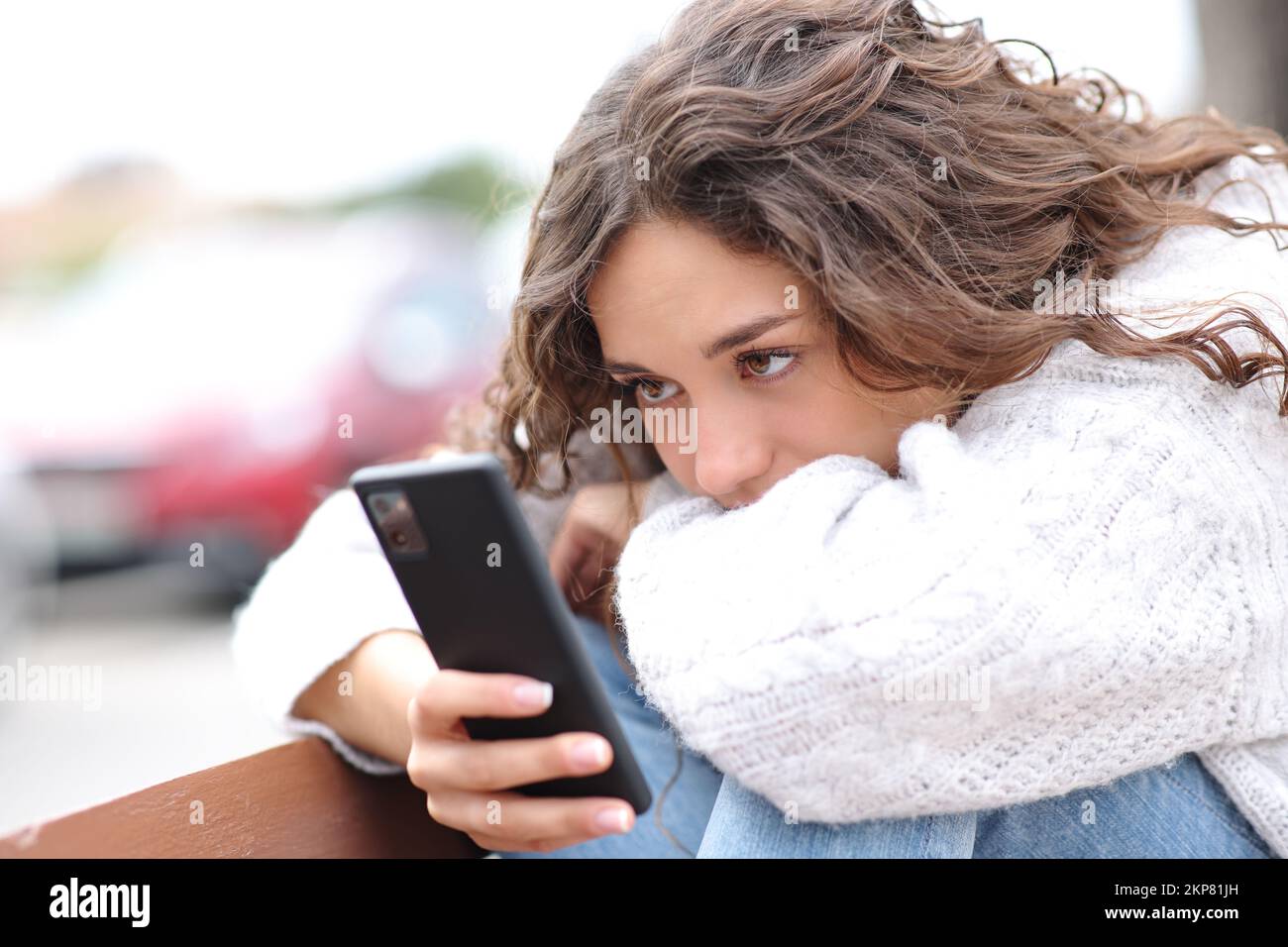 Donna triste che controlla lo smartphone seduto in una panca per strada Foto Stock