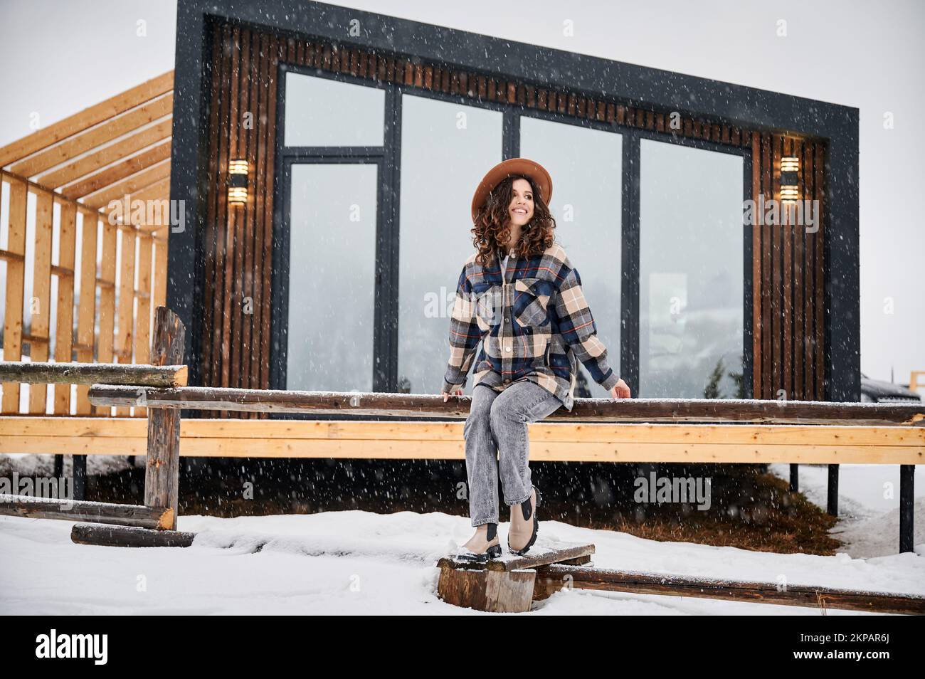 Giovane donna curly che riposa sulla terrazza della moderna casa di fienile in montagna. Felice turista femmina godendo le vacanze invernali in nuovo cottage. Foto Stock