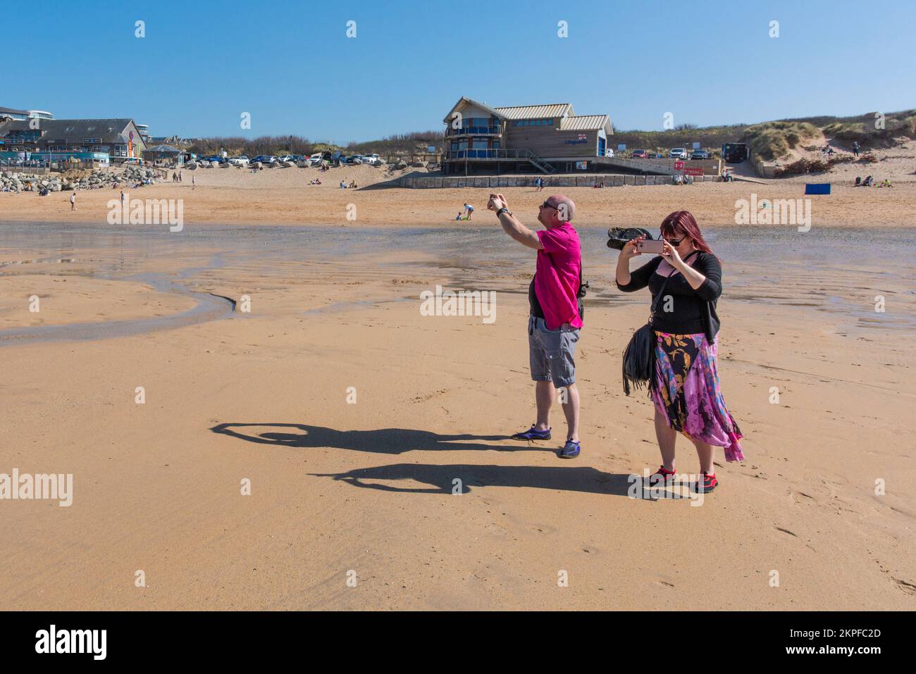 Ombre lanciate da due vacanzieri che scattano fotografie con i loro telefoni cellulari su Fistral Beach a Newquay in Cornovaglia nel Regno Unito. Foto Stock