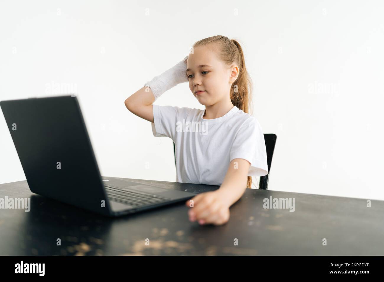 Preoccupata bambina bionda mettere mano rotta avvolto in cerotto bianco sulla testa pensieroso guardando lo schermo del notebook seduto al tavolo su bianco Foto Stock