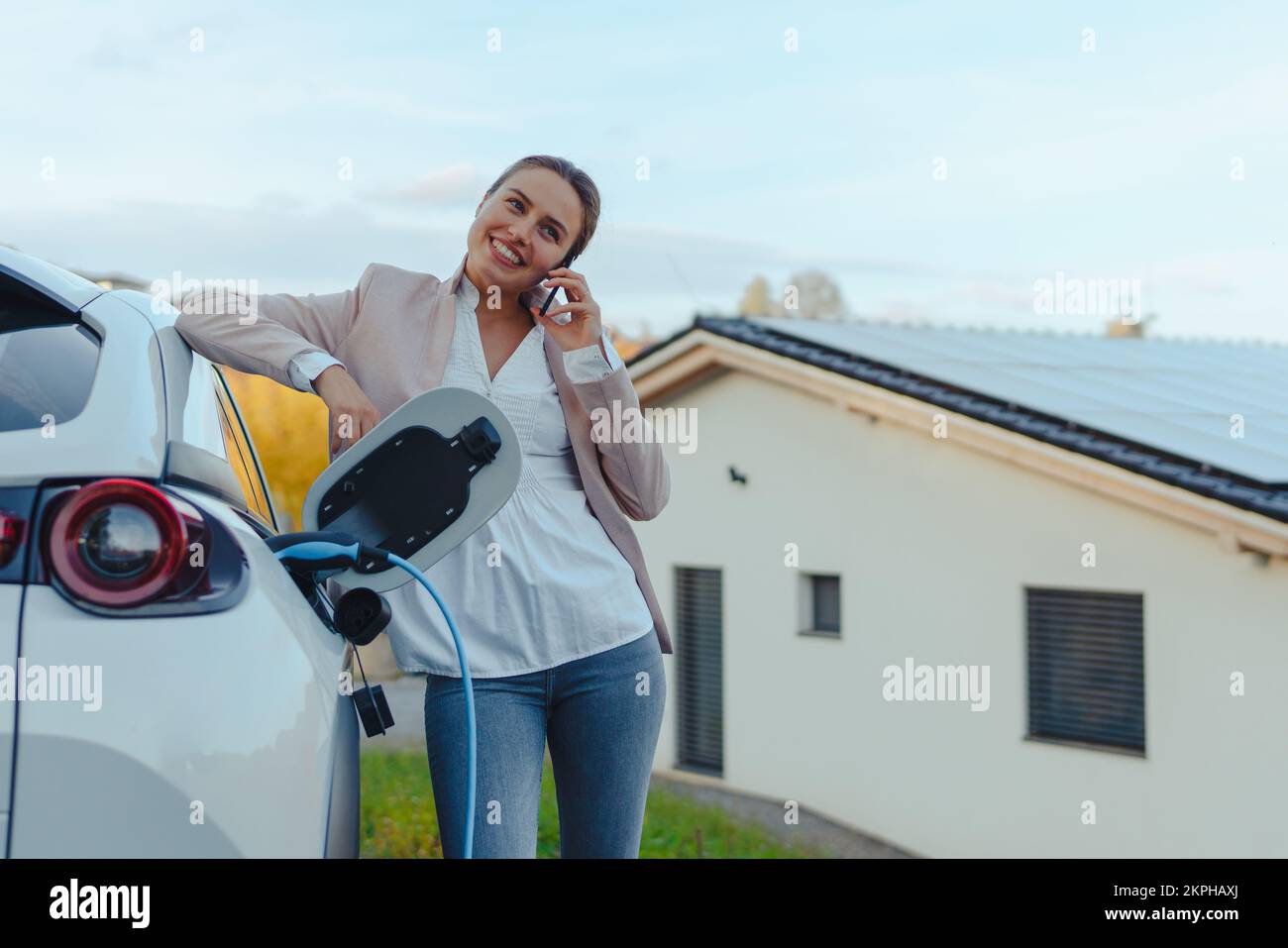 Giovane donna con smartphone in attesa mentre la sua auto elettrica in carica nella stazione di ricarica a casa, concetto di trasporto sostenibile ed economico. Foto Stock