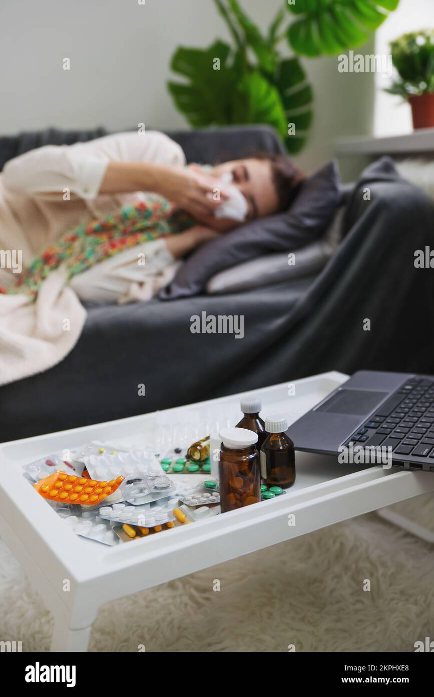 Una giovane donna malata con un virus, naso che cola o reazione allergica si trova su un divano a casa con focus su vari farmaci e pillole. Concetto di pandemia virale. Foto Stock