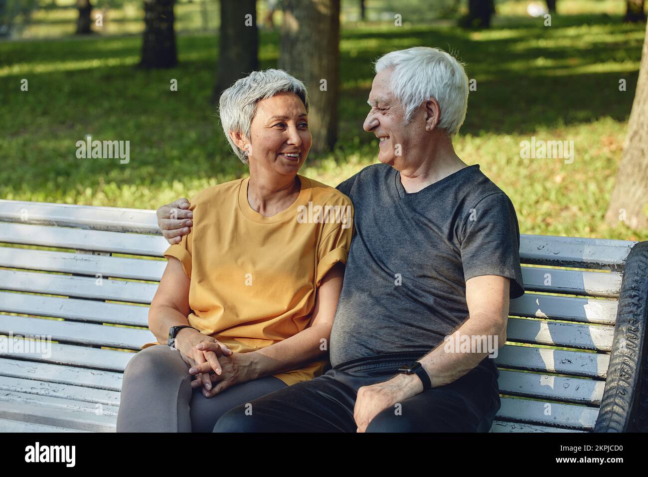 Felice multi razziale anziano coppia di capelli grigi riposati sedersi sulla panca nel parco dopo una passeggiata, mattina a piedi sorridendo godere di relax all'aperto durante l'estate soleggiata Foto Stock