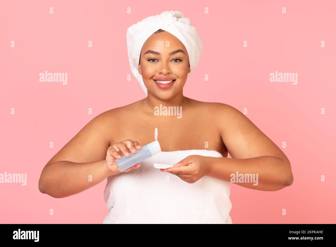 Giovane donna nera corpo positivo versando acqua micellare al tampone di cotone per la pulizia della pelle, in piedi avvolto in asciugamano da bagno Foto Stock