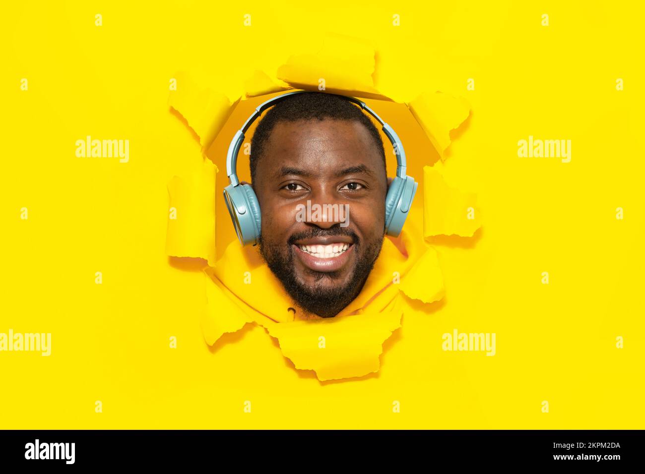 Felice uomo afroamericano che indossa cuffie wireless e sorridente, ascoltando la musica attraverso il buco in carta gialla strappata Foto Stock