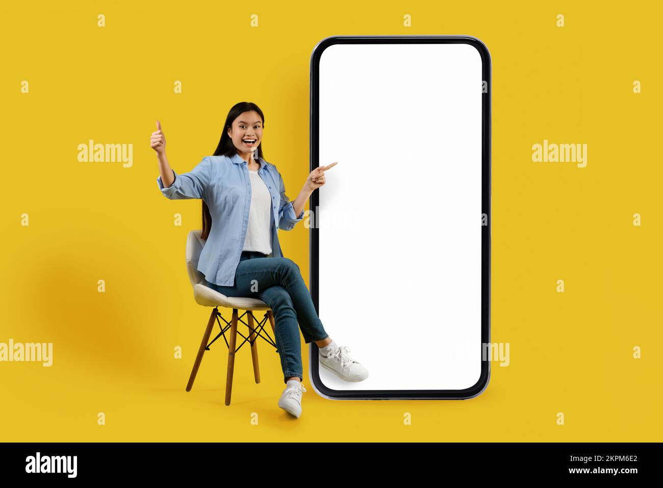 Felice scioccato millenario cinese lady studente in casual sedersi sulla sedia, punta le dita a grande smartphone con schermo vuoto Foto Stock
