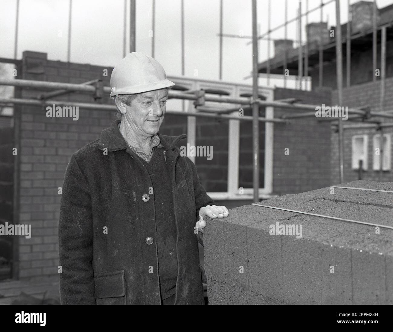 1980s, in un cantiere di costruzione di alloggi, un caposquadra in una giacca da asino e un elmetto che controlla una pila di blocchi da costruzione in cemento leggero, utilizzati nella costruzione di nuove case, Inghilterra, Regno Unito. Foto Stock