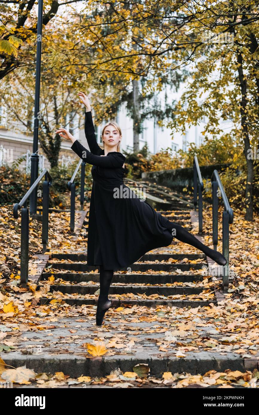 Ballerina Dancing in via autunnale città, ballerino moderno in vestito nero, Pointe Shoes Outdoors. Balsamo, balsamo quotidiano Foto Stock
