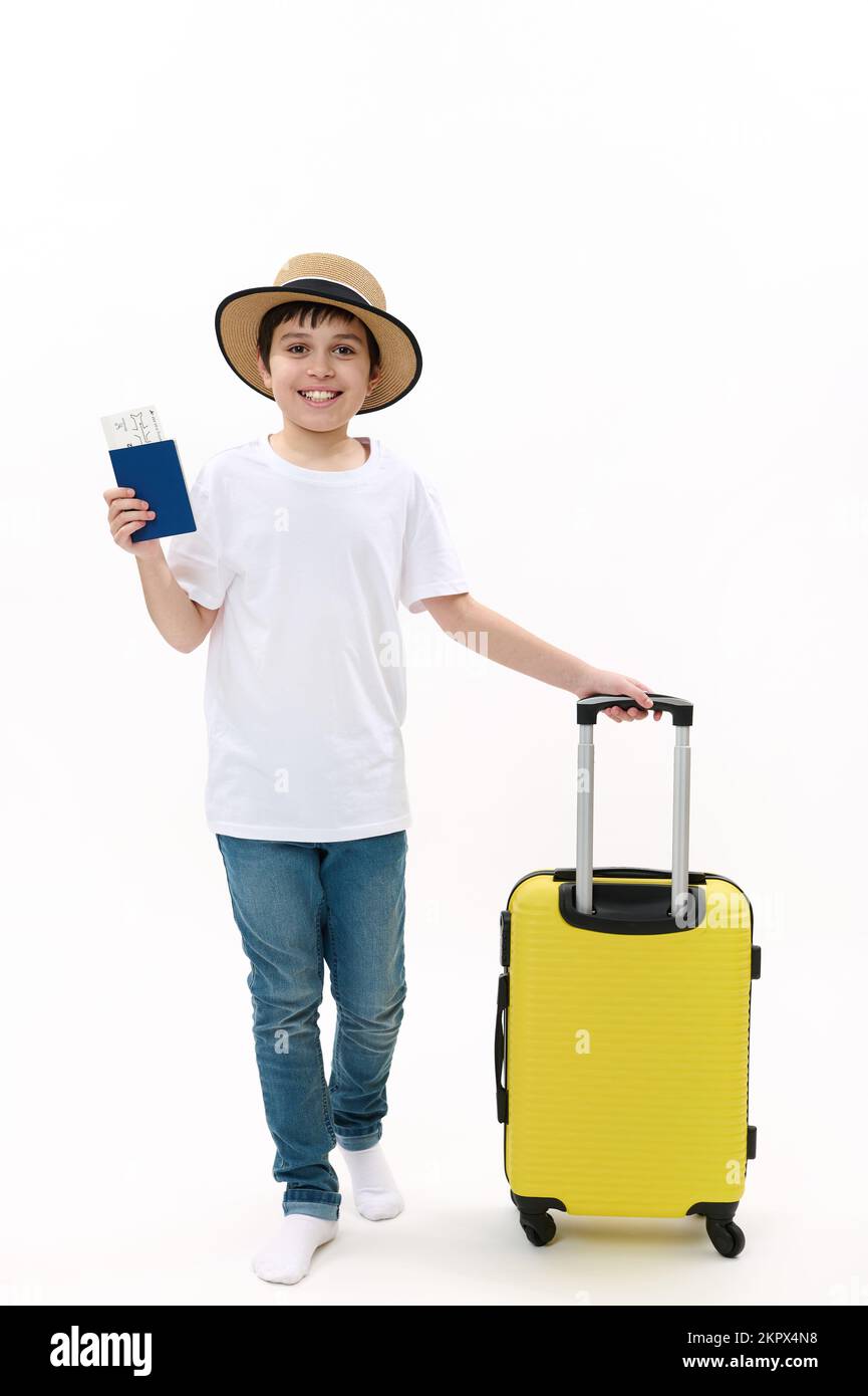 Ragazzo viaggiatore adolescente in t-shirt casual, jeans e cappello di paglia, passeggiate con carta d'imbarco e valigia su sfondo bianco. Foto Stock