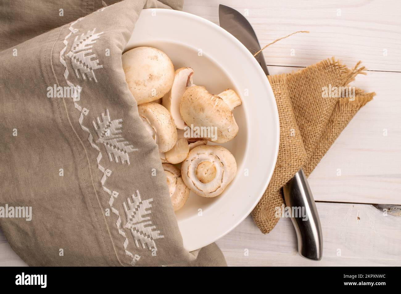 Diversi funghi freschi e biologici champignon appetitosi in un piatto di ceramica bianca, con un coltello di metallo e due tovaglioli di lino e iuta, su un tavolo. Foto Stock
