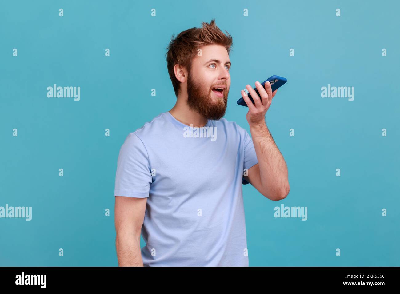 Ritratto di un uomo ottimista che registra un messaggio vocale o parla con un assistente online sul suo smartphone tenendo il telefono vicino alla bocca. Studio in interni isolato su sfondo blu. Foto Stock
