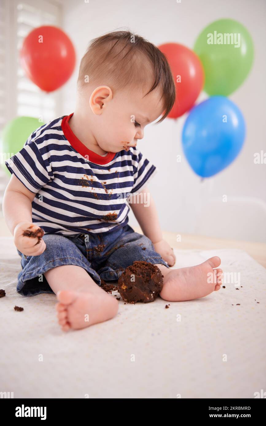 HES assaggiando la sua torta di compleanno. un bambino seduto a casa con palloncini sullo sfondo. Foto Stock