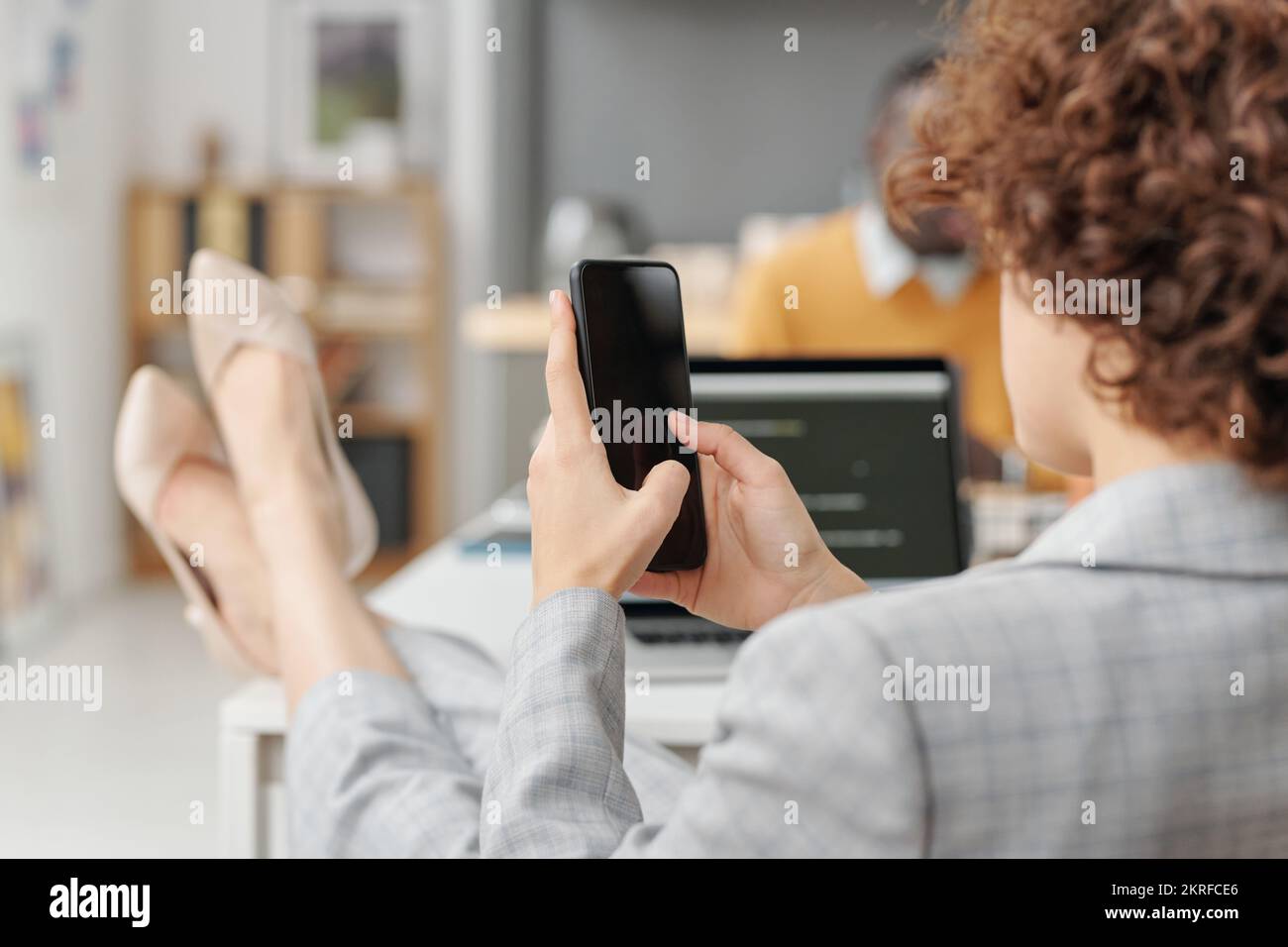 Vista posteriore della giovane donna d'affari che tocca lo schermo del suo smartphone mentre si riposa dopo il lavoro in ufficio Foto Stock