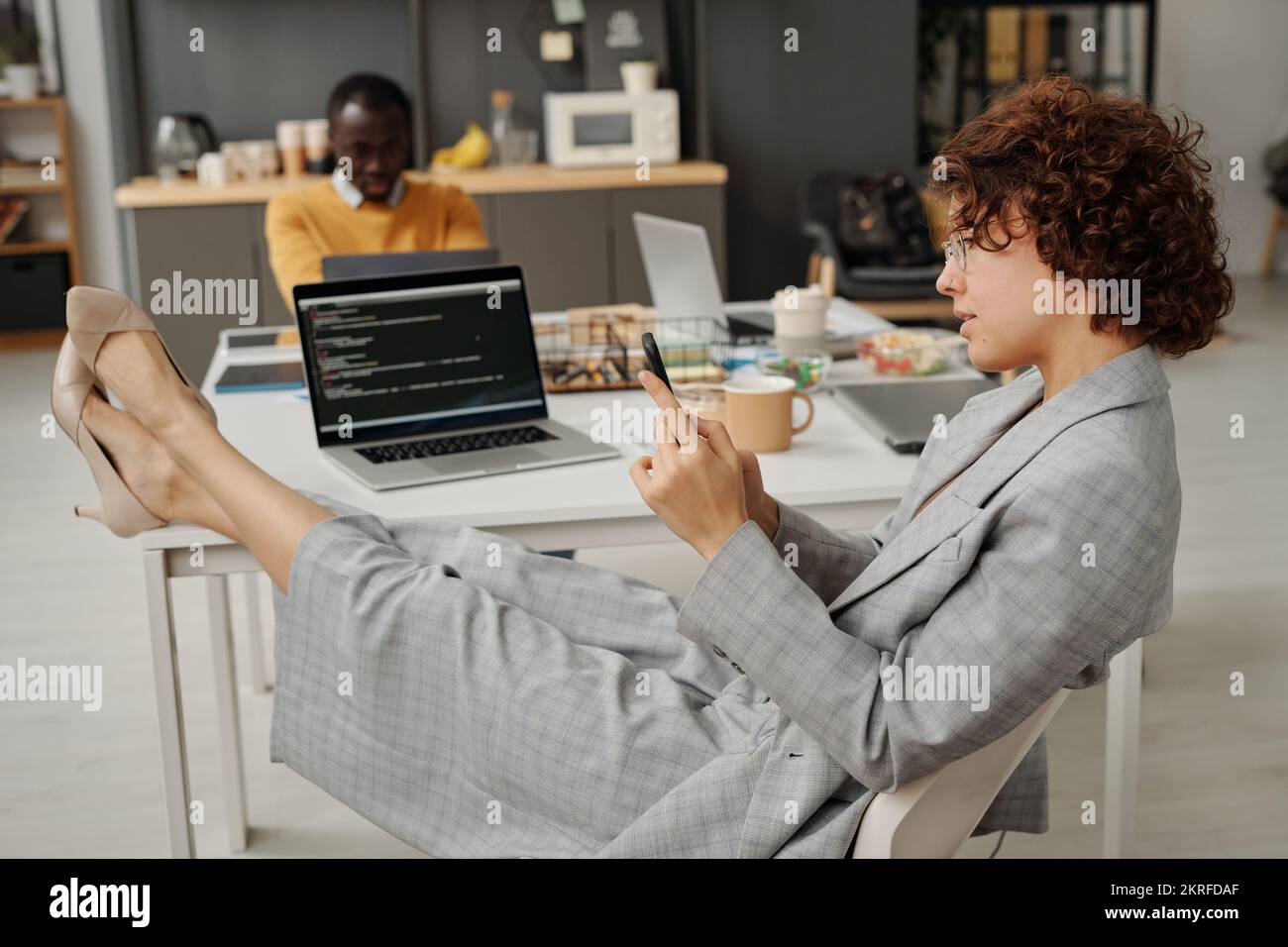 Giovane donna d'affari seduta sul posto di lavoro con un notebook e che usa lo smartphone durante la pausa in ufficio Foto Stock