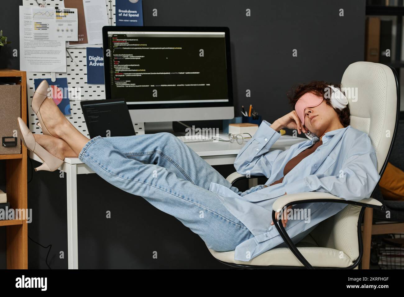 Giovane donna d'affari in maschera che dorme sul suo posto di lavoro durante la giornata lavorativa in ufficio Foto Stock