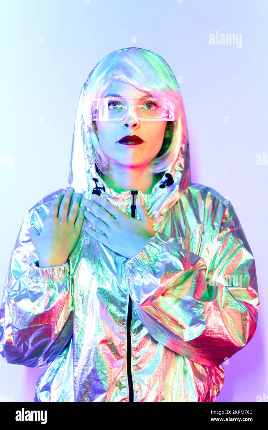 Giovane donna in parrucca con occhiali intelligenti per realtà aumentata con schermo virtuale che visualizza icone mentre si è in piedi in studio con luci blu al neon incandescenti Foto Stock