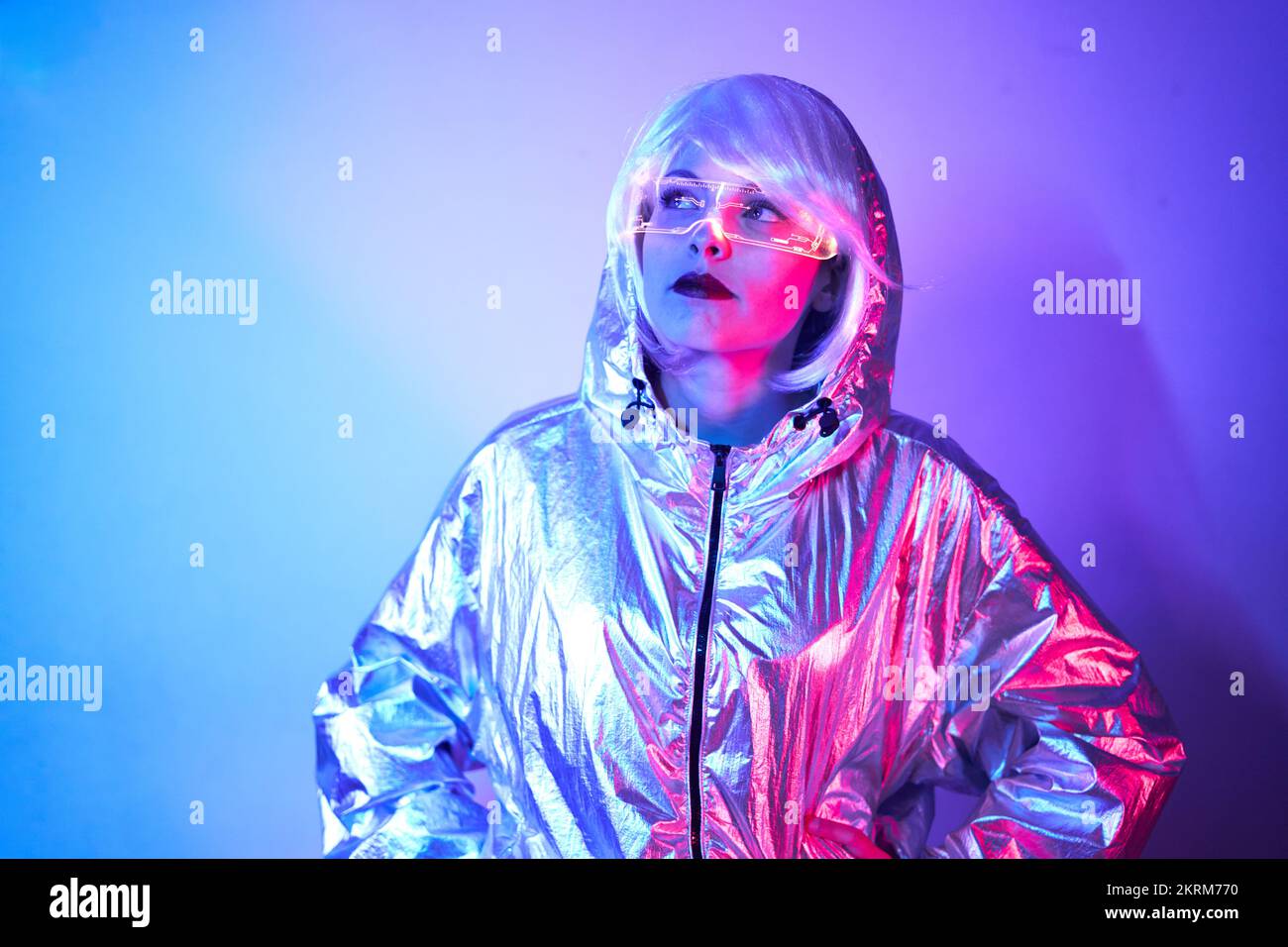 Giovane donna in parrucca con occhiali intelligenti per realtà aumentata con schermo virtuale che visualizza icone mentre si è in piedi in studio con luci blu al neon incandescenti Foto Stock
