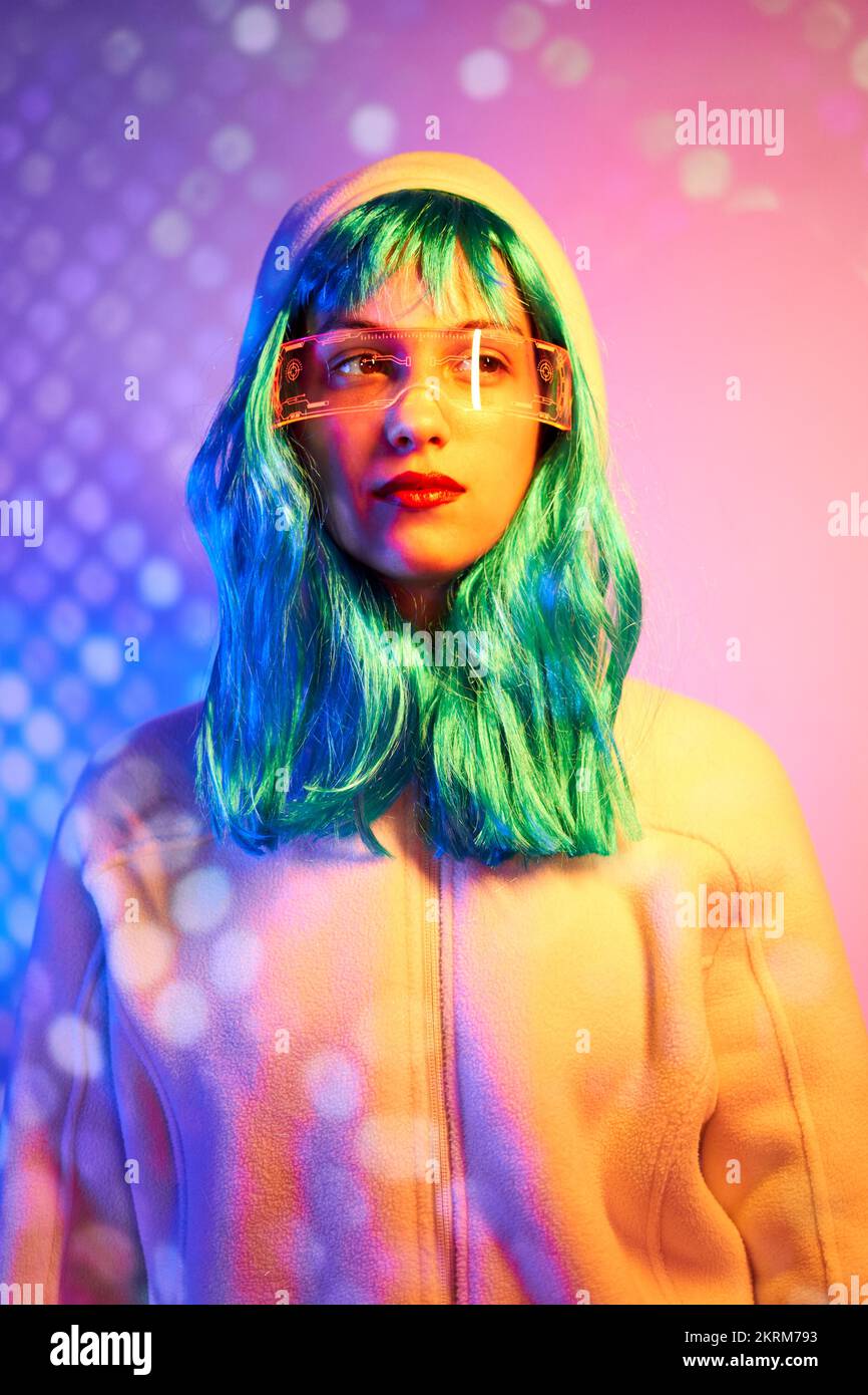 Giovane donna in parrucca con occhiali intelligenti per realtà aumentata con schermo virtuale che visualizza icone mentre si è in piedi in studio con luci rosa al neon scintillanti Foto Stock