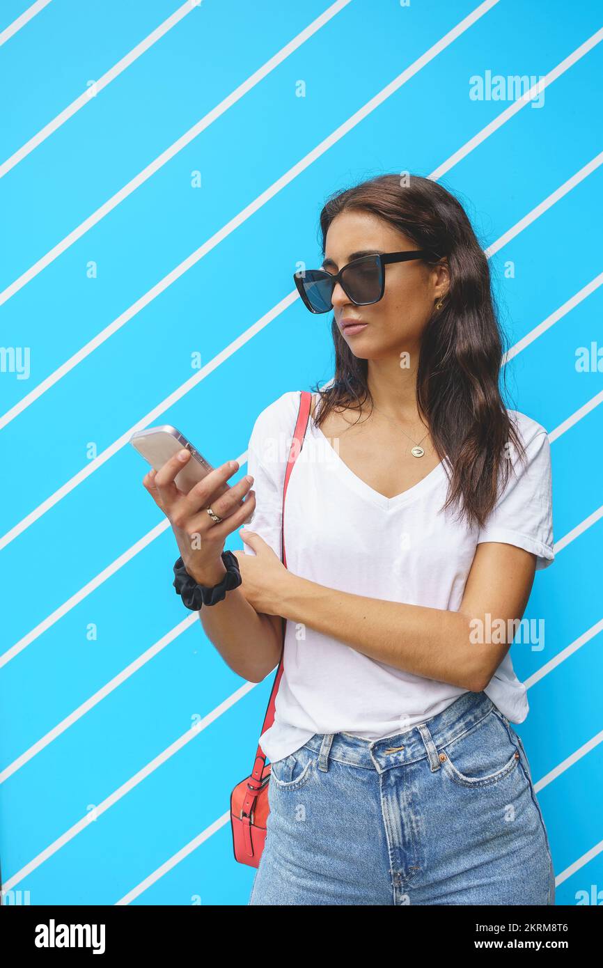 Donna in T-shirt bianca e jeans guardando lo schermo del telefono cellulare, mentre navigando su Internet su sfondo a strisce blu Foto Stock