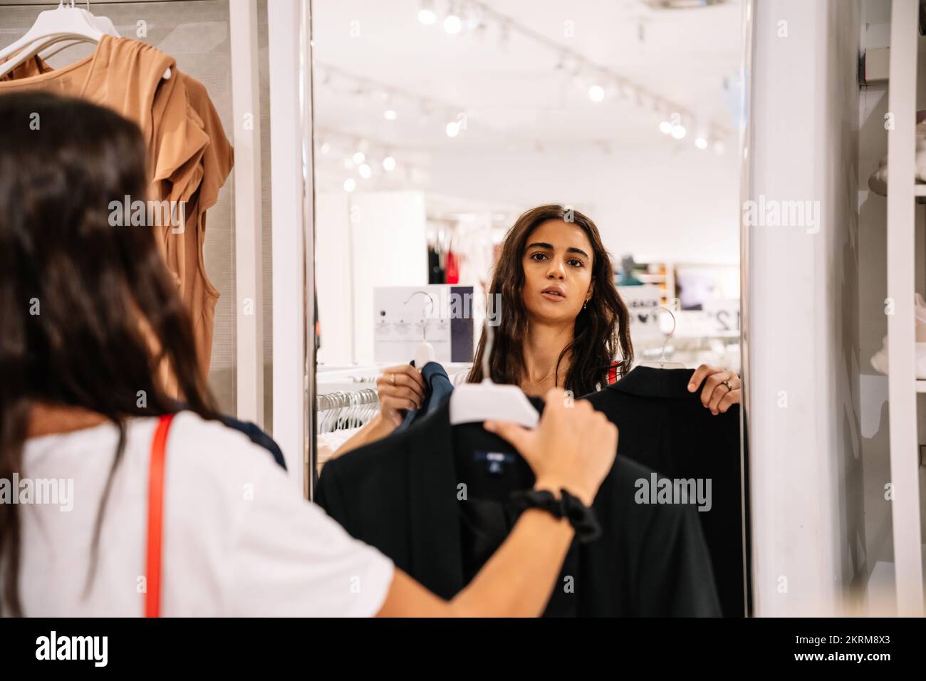 Giovane cruna che guarda lo specchio mentre seleziona nero elegante giacca su grucce nel mercato moderno Foto Stock