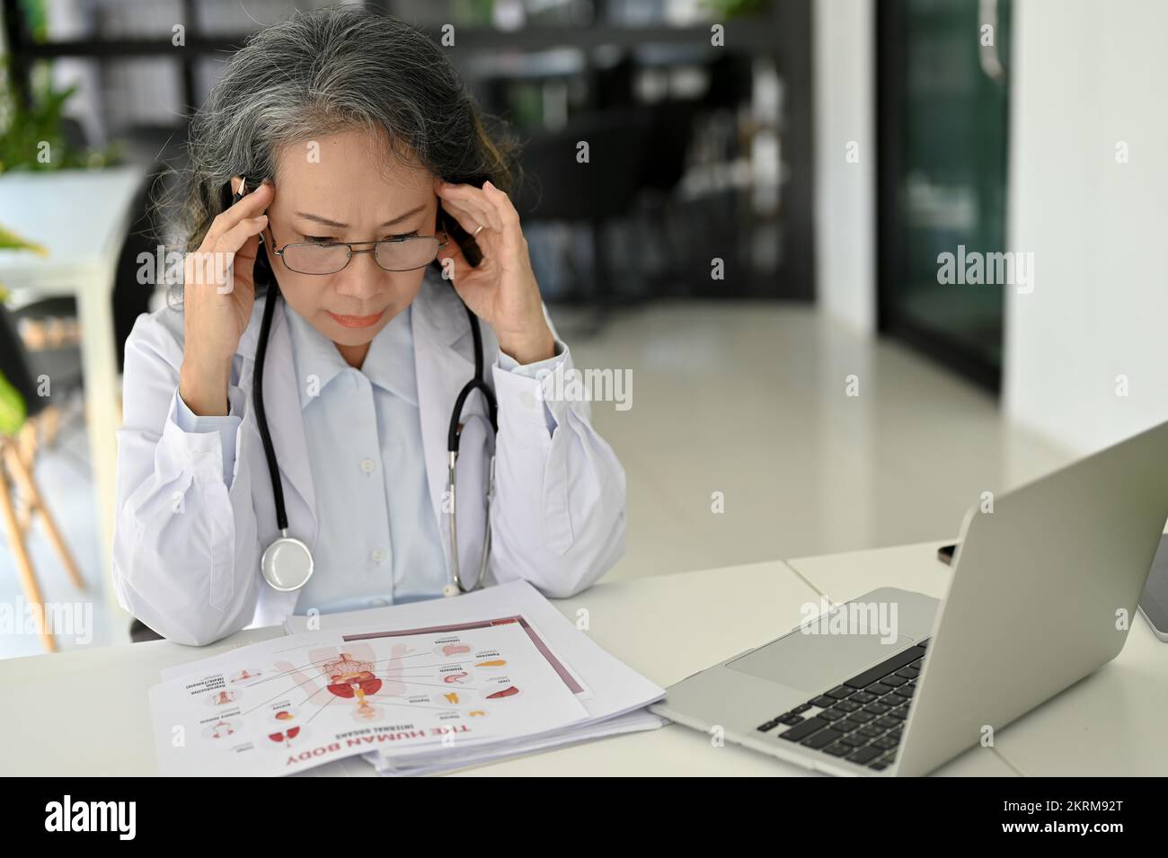 Stressato e professionale asiatico anziano medico concentrandosi sul suo caso di ricerca medica e la lettura di carta di ricerca alla sua scrivania. Foto Stock
