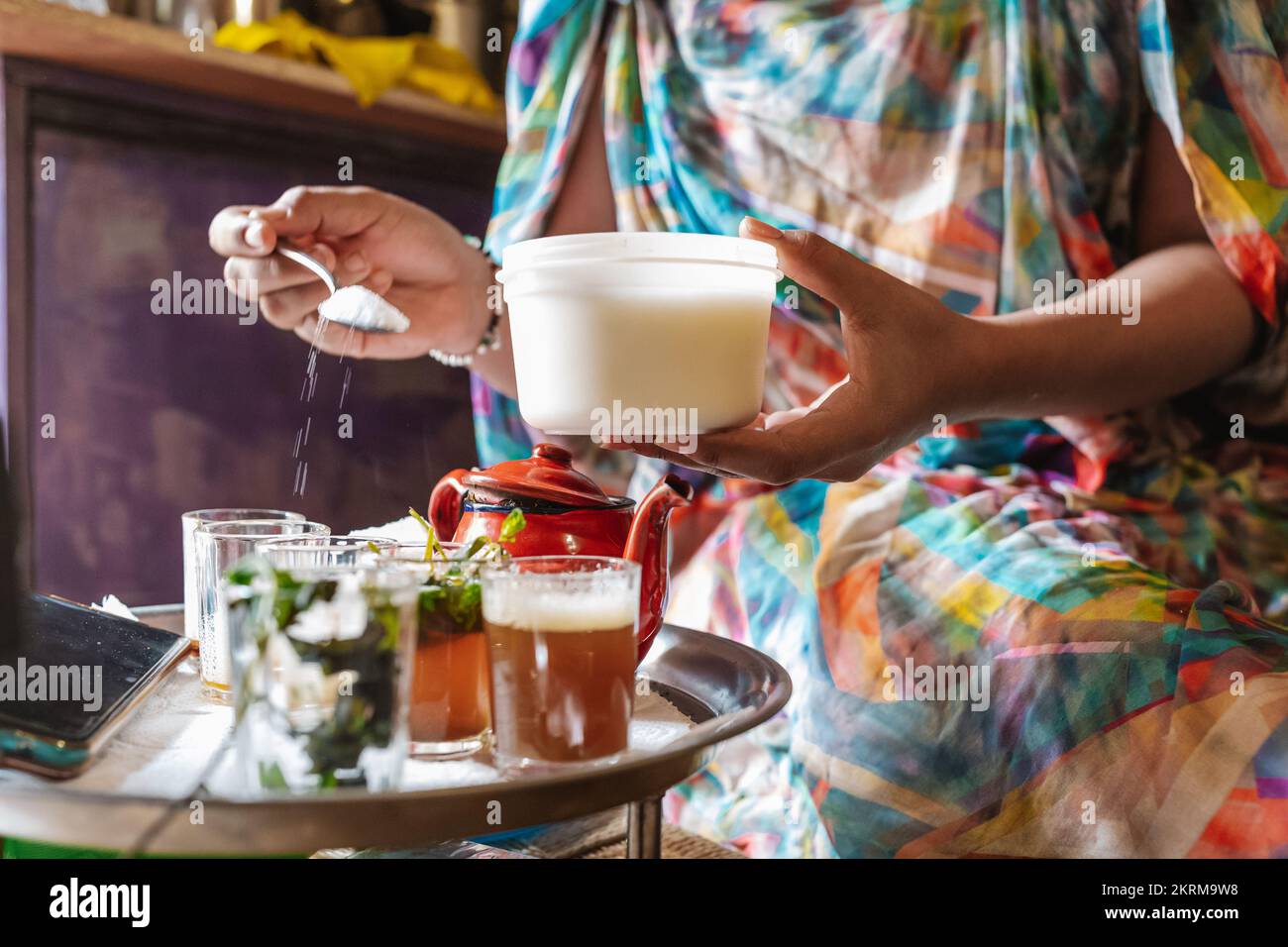 Crop femmina in abito con forme geometriche aggiungendo zucchero nelle bevande con erbe poste sul tavolo da caffè rotondo con teiera Foto Stock