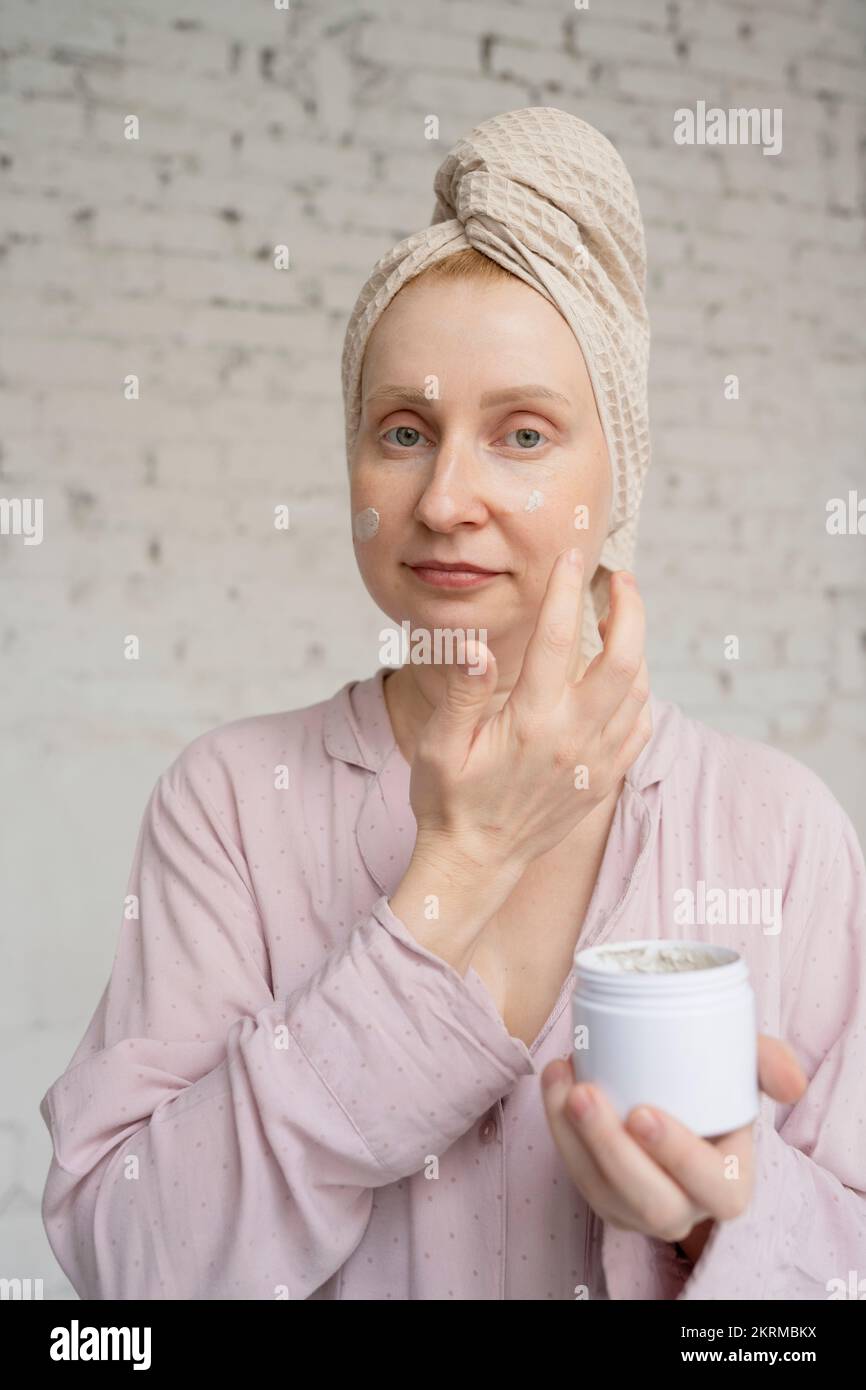 Contenuto femmina con asciugamano sulla testa e vasetto di crema che ha la procedura di cura della pelle mentre in piedi in bagno chiaro contro muro di mattoni Foto Stock
