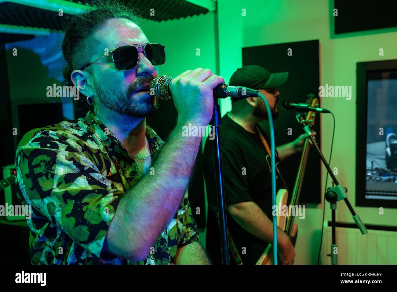 Uomo che suona la chitarra elettrica mentre il cantante canta la canzone nel microfono durante le prove in studio scuro con illuminazione al neon brillante Foto Stock