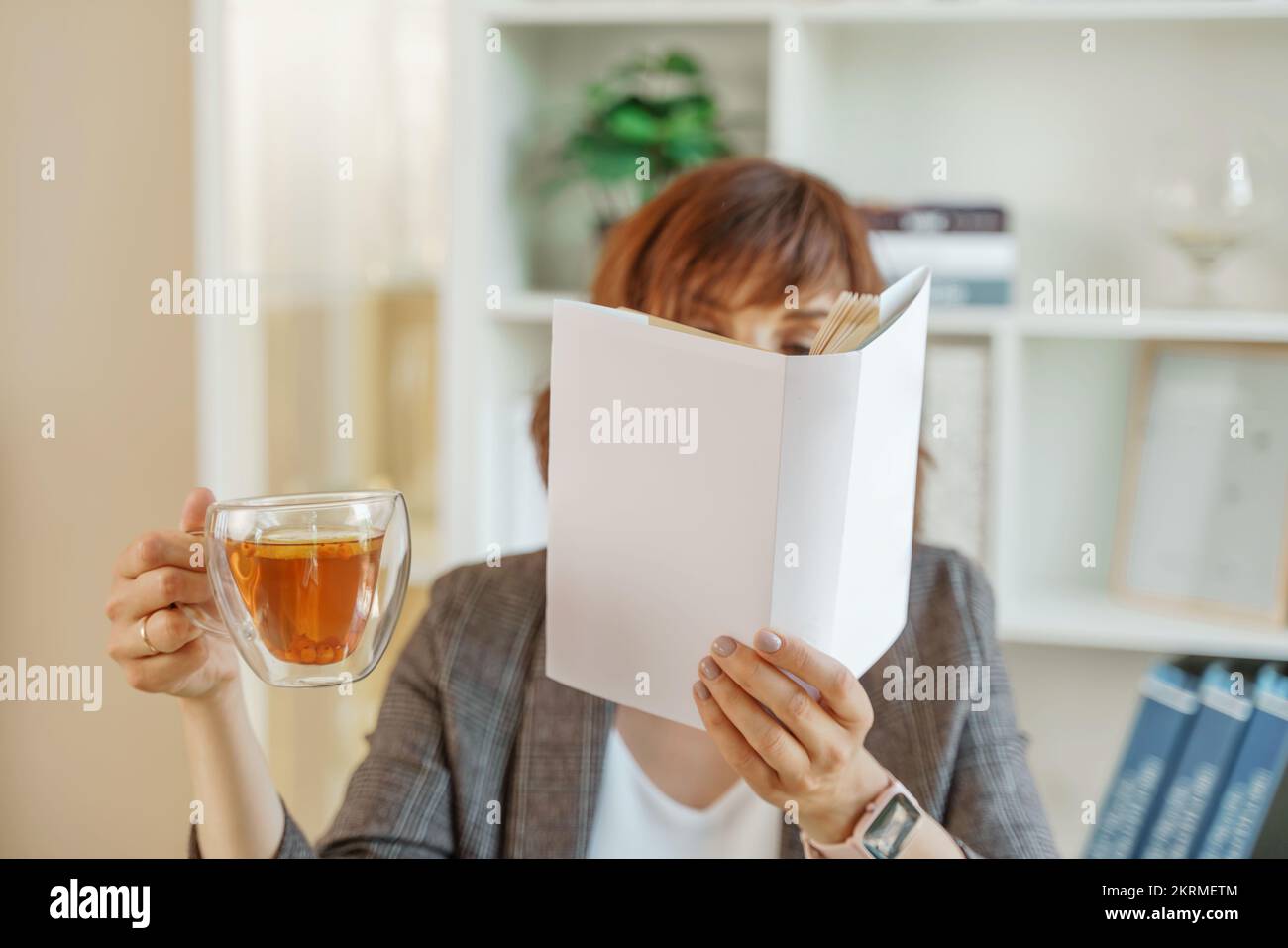 Ritratto di una ragazza che tiene un libro aperto al suo volto in una mano e una tazza di tè nell'altra sullo sfondo dell'interno. Foto Stock