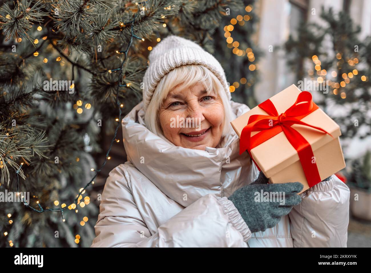 Buon Natale e buone feste. Donna anziana che si diverte con una scatola regalo a sorpresa presente vicino all'albero di Natale all'aperto al mercatino di Natale in città Foto Stock