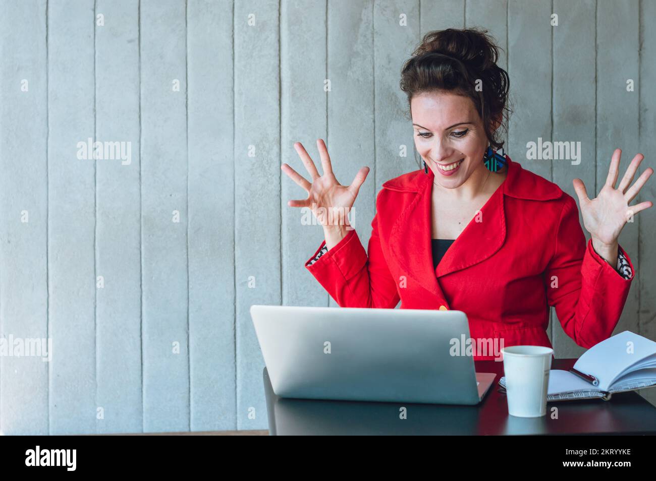 Donna adulta caucasica che reagisce felicemente con le mani sollevate e aperte, sorridendo mentre lavora sul suo computer portatile, seduto in una caffetteria. Foto Stock