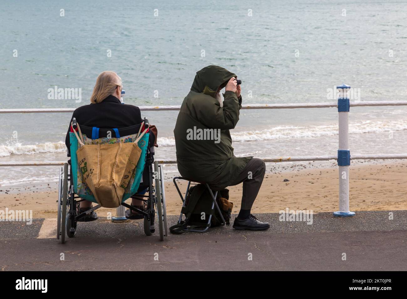 Coppia seduta sulla passeggiata lungomare, donna in sedia a rotelle, uomo in sedile pieghevole con fotocamera tenuto a scattare foto, a Weymouth, Dorset UK nel mese di ottobre Foto Stock