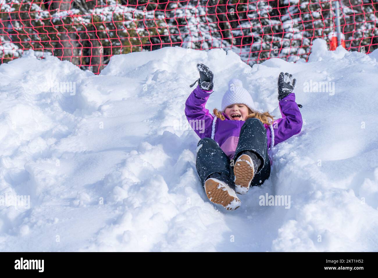 Una bella ragazza in un vestito da sci viola scivola giù dalla collina di neve. Concetto di giochi per bambini nel periodo invernale. Foto di alta qualità Foto Stock