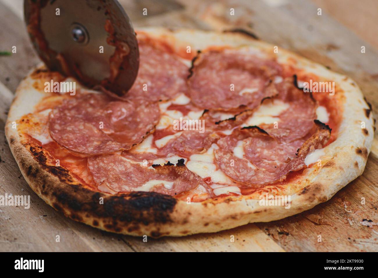 Tagliare la pizza calda con salsa di pomodoro, mozzarella e salame con un taglierino su un asse di legno, affumicato, primo piano Foto Stock