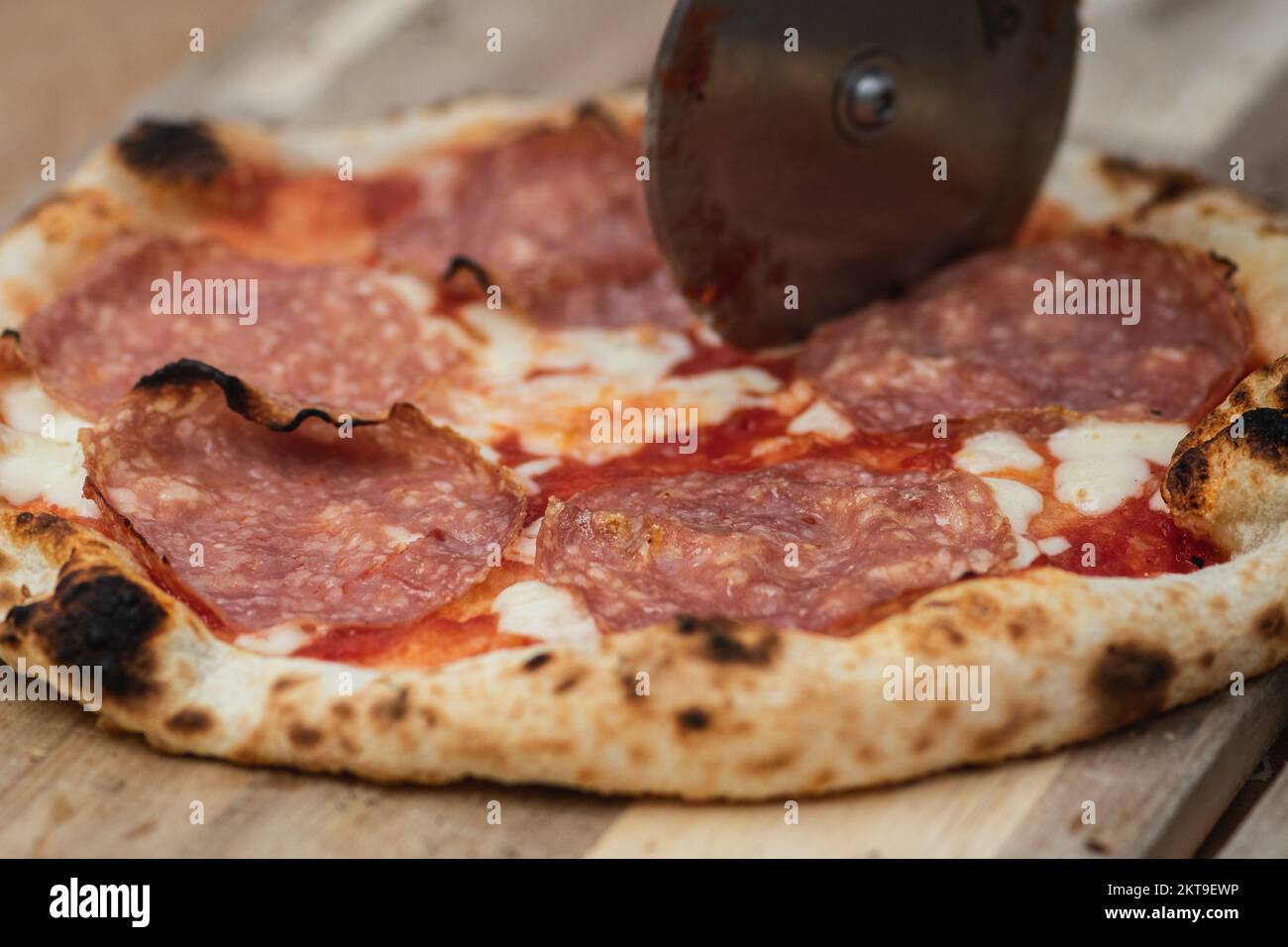 Tagliare la pizza calda con salsa di pomodoro, mozzarella e salame con un taglierino su un asse di legno, affumicato, primo piano Foto Stock