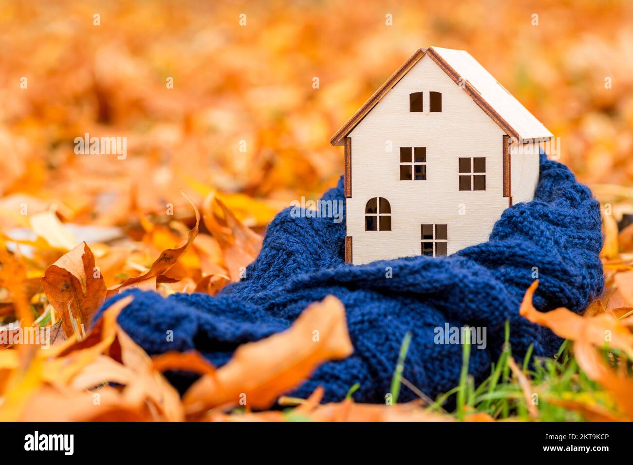 La casa di legno giocattolo è avvolta in una sciarpa calda sullo sfondo delle foglie autunnali Foto Stock
