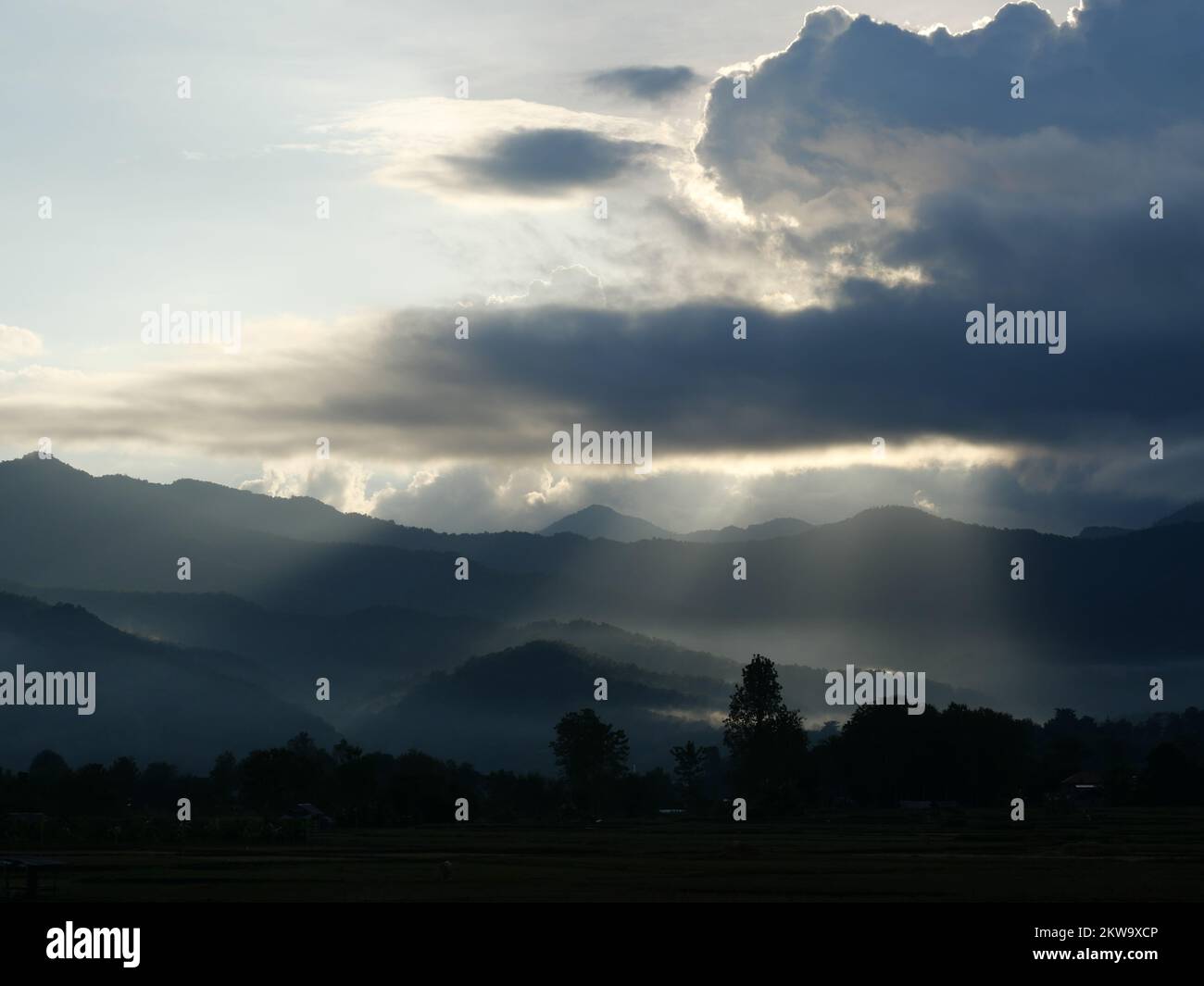 Silhouette di albero e montagna con luce solare sparare attraverso la nuvola scura alla terra all'alba, Mist copre la foresta e le montagne all'alba Foto Stock