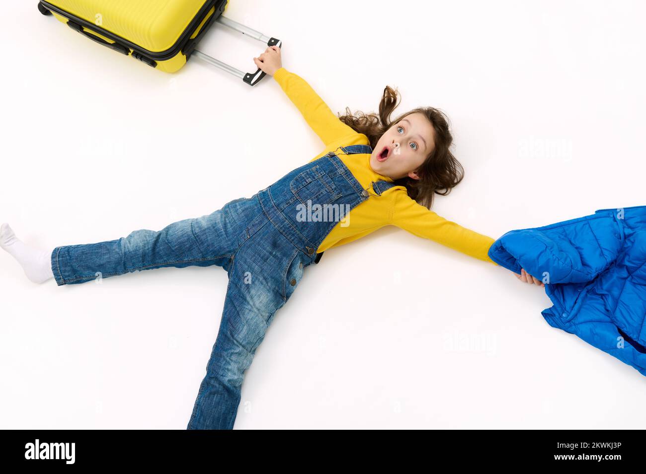 La bambina carina tiene il suo cappotto e la sua valigia in mani tirate, esprimendo emozione, stupore e stupore, Foto Stock