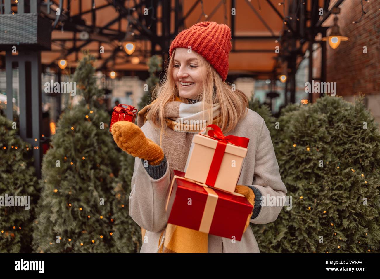Donna bionda felice in cappotto di inverno che tiene una scatola regalo al mercato di Natale. Attraente ragazza felice shopper tenere scatole regalo, borse di shopping, canna caramella in Foto Stock