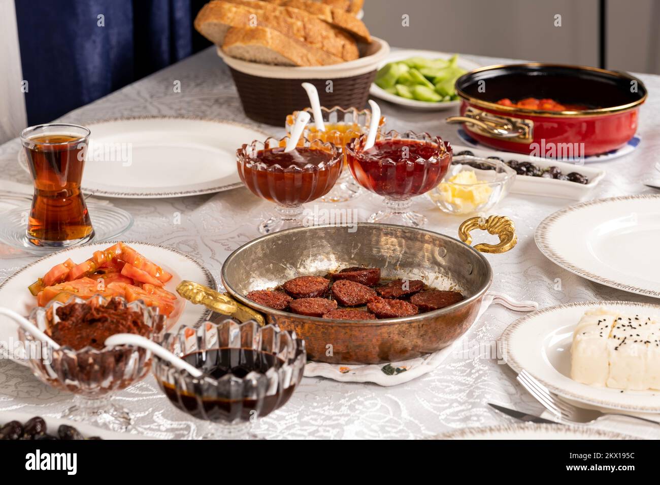 Mattina routine ricco tavolo per la colazione. Salsiccia turca chiamata sucuk in padella pronta per mangiare. Pane, formaggio, pomodori, marmellata, miele, tè in vetro. Foto Stock