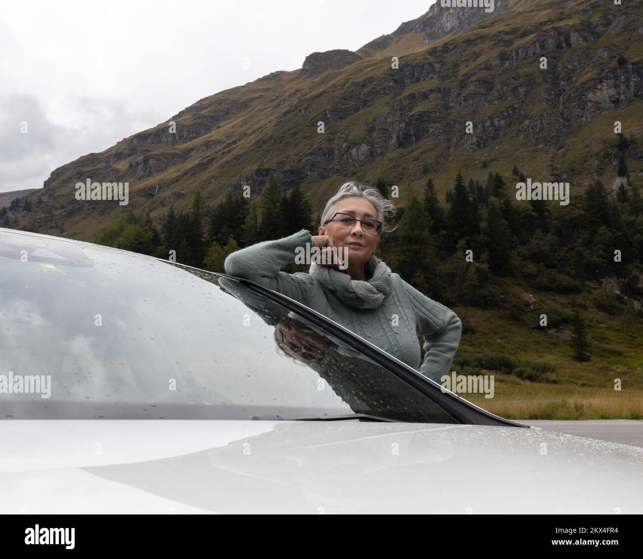 Una giovane donna dai capelli grigi in un caldo maglione si trova vicino ad un'auto in una valle alpina Foto Stock