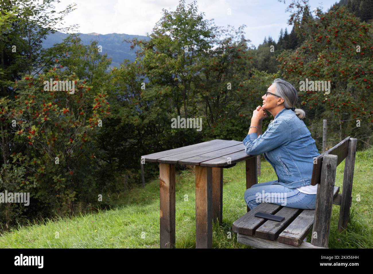 Una donna dai capelli grigi siede su un tavolo di legno, in un ambiente alpino. Foto Stock