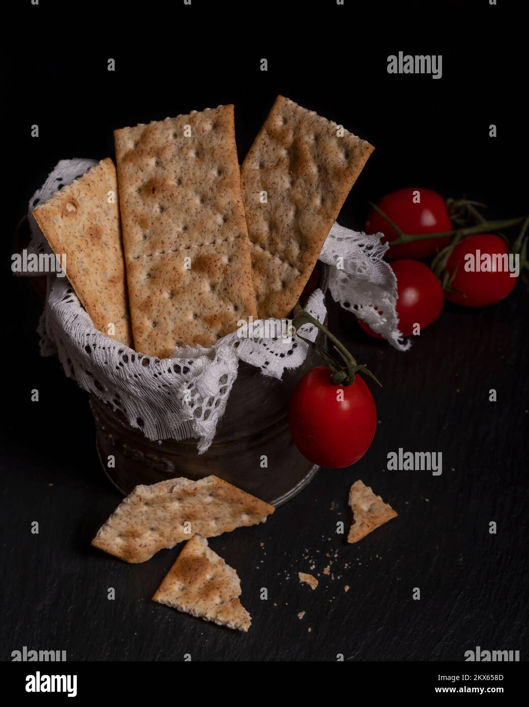 I cracker sono in un contenitore di latta, con i pomodori ciliegini vicini, su uno sfondo nero Foto Stock