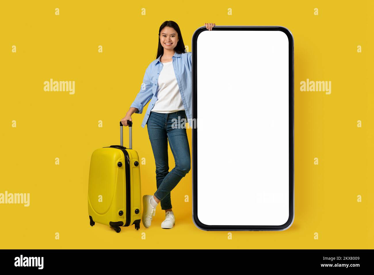 Sorridente giovane studentessa coreana in un casual con valigie con enorme smartphone Foto Stock
