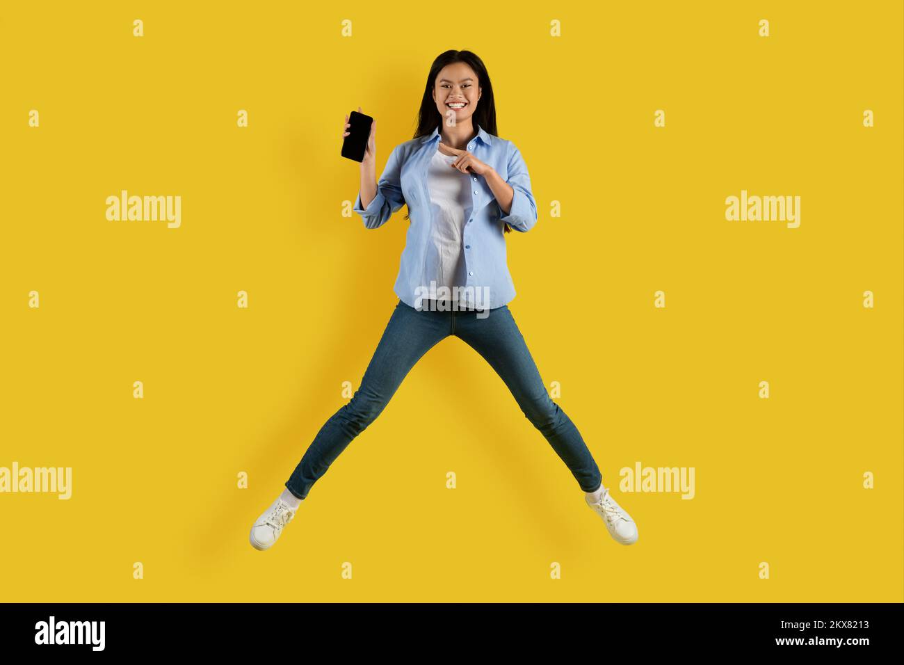 Soddisfatto simpatico studente asiatico millennial salto, congelare in aria, puntare il dito contro lo smartphone con schermo vuoto Foto Stock
