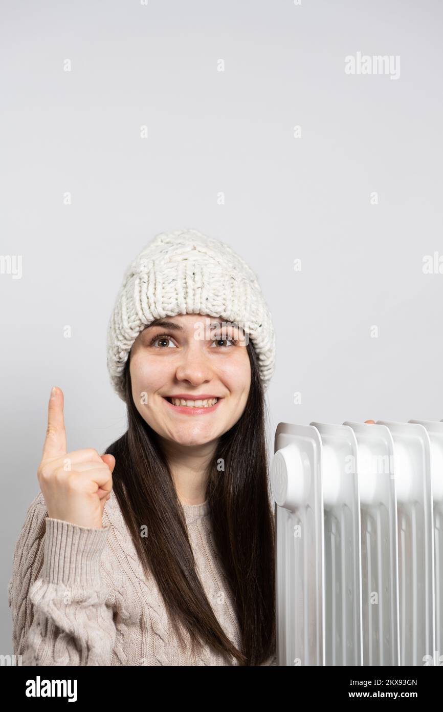 Una donna in un cappello che si scalda vicino al riscaldatore dell'olio e guarda e punta con il dito. Foto Stock