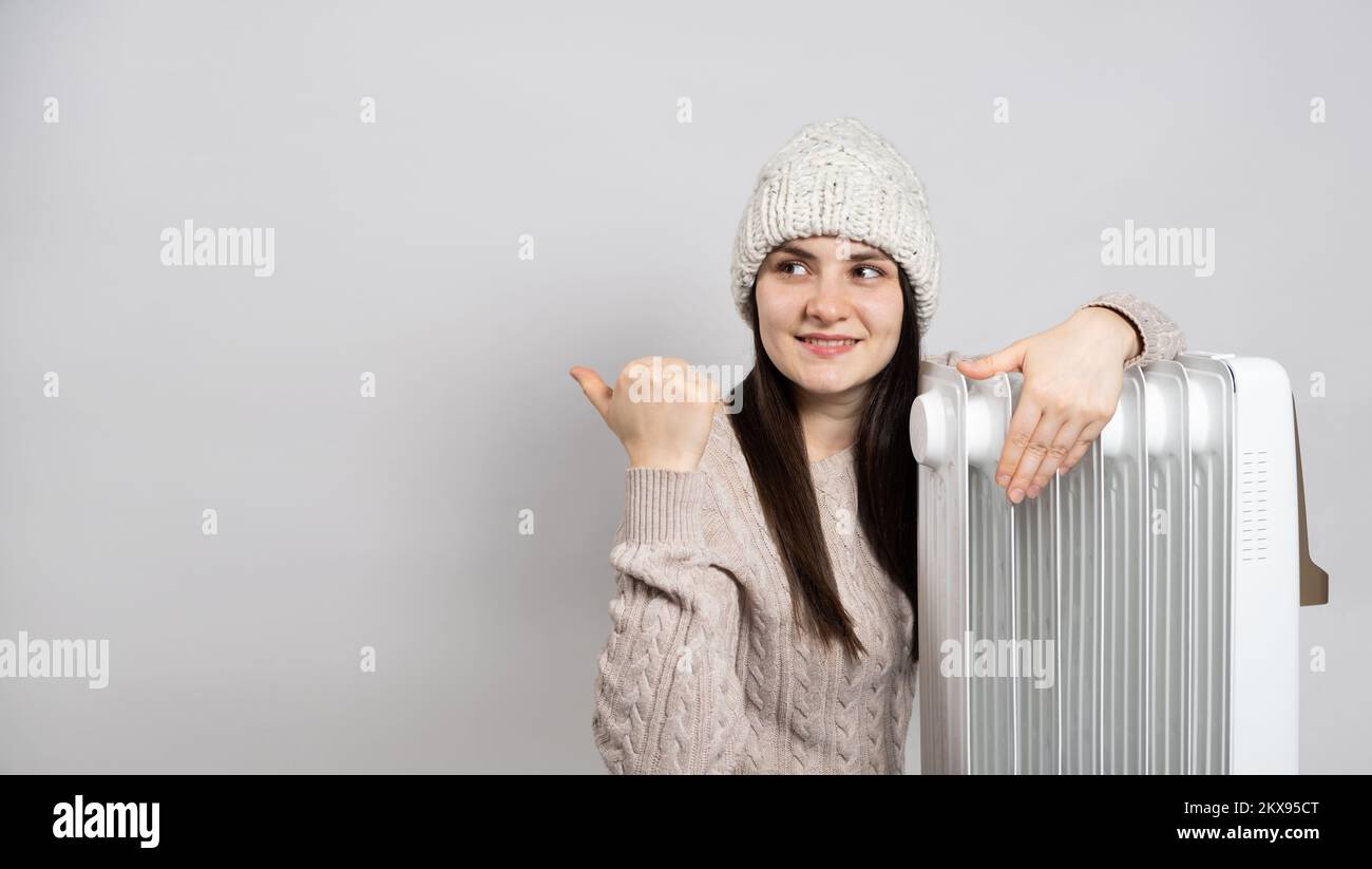 Una donna in un cappello abbraccia un riscaldatore di olio e punta il suo dito al lato sul posto per il testo. Foto Stock