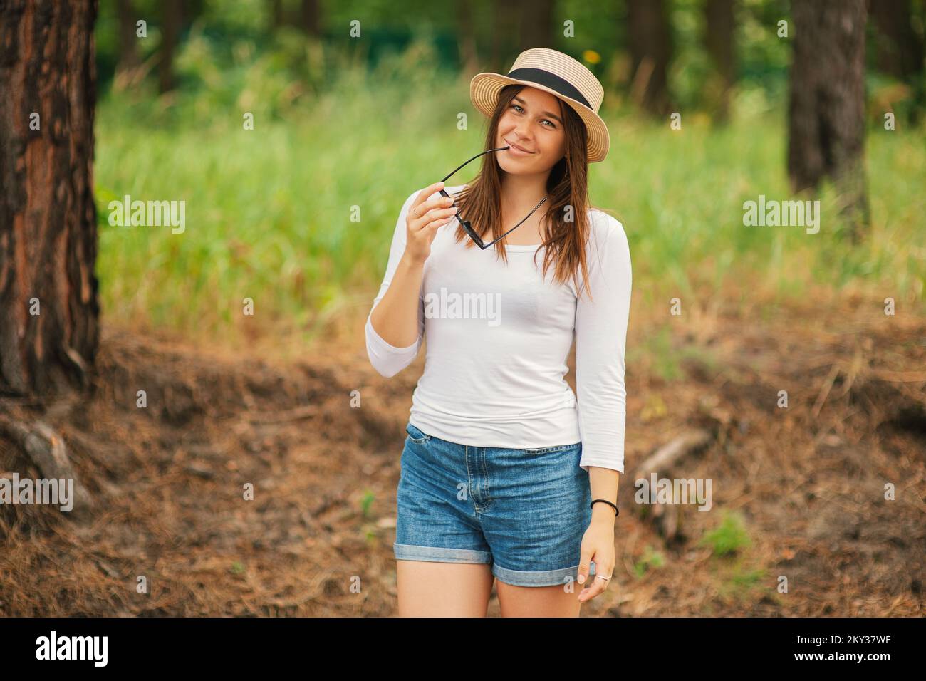 Carino giovane donna alla moda indossando un cappello di regolazione di estate tenendo gli occhiali da sole nelle sue mani a guardare la macchina fotografica in piedi all'aperto Foto Stock