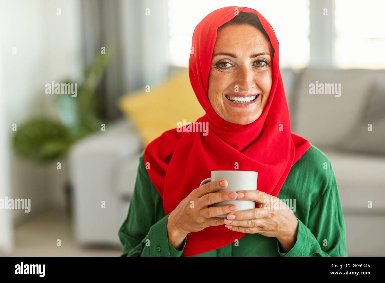 Ritratto di felice donna islamica di mezza età in hijab seduta in soggiorno con tazza di tè alle erbe e sorridente alla macchina fotografica Foto Stock